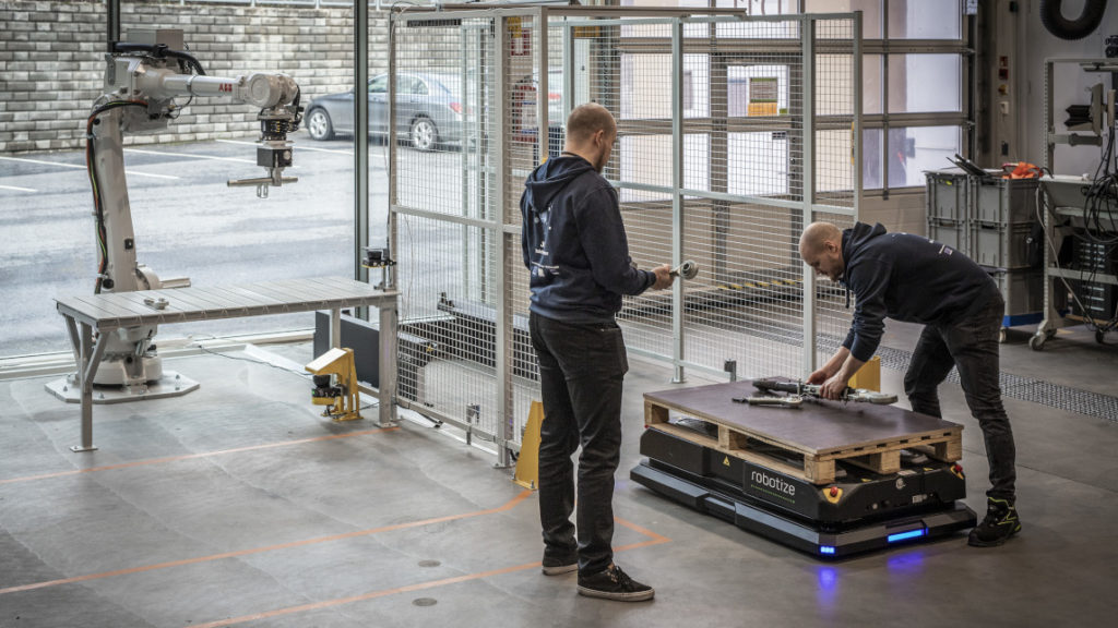 Kaksi miestä tuotantotiloissa ja lattialla matala robotti, jonka päällä on vaihtolava.